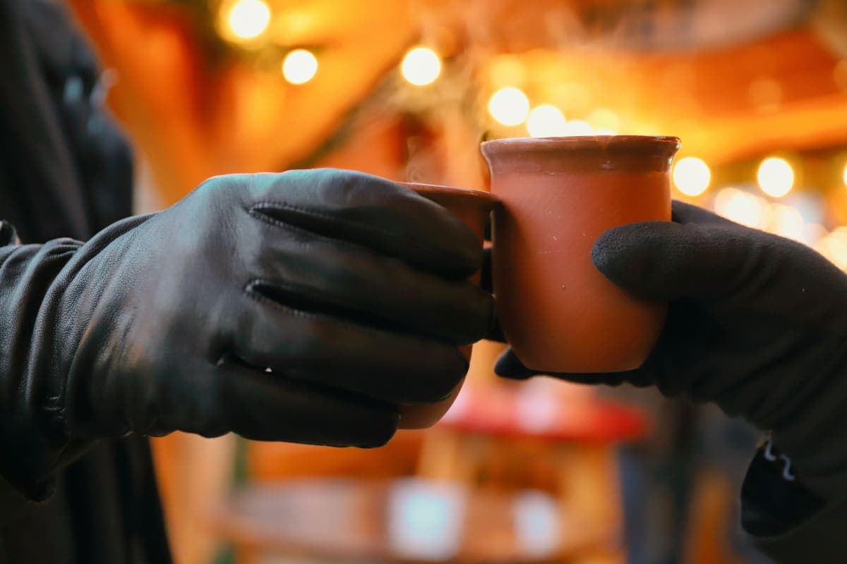Two people clinking mugs of mulled wine at an evening Christmas market stall, with market lights glowing behind them