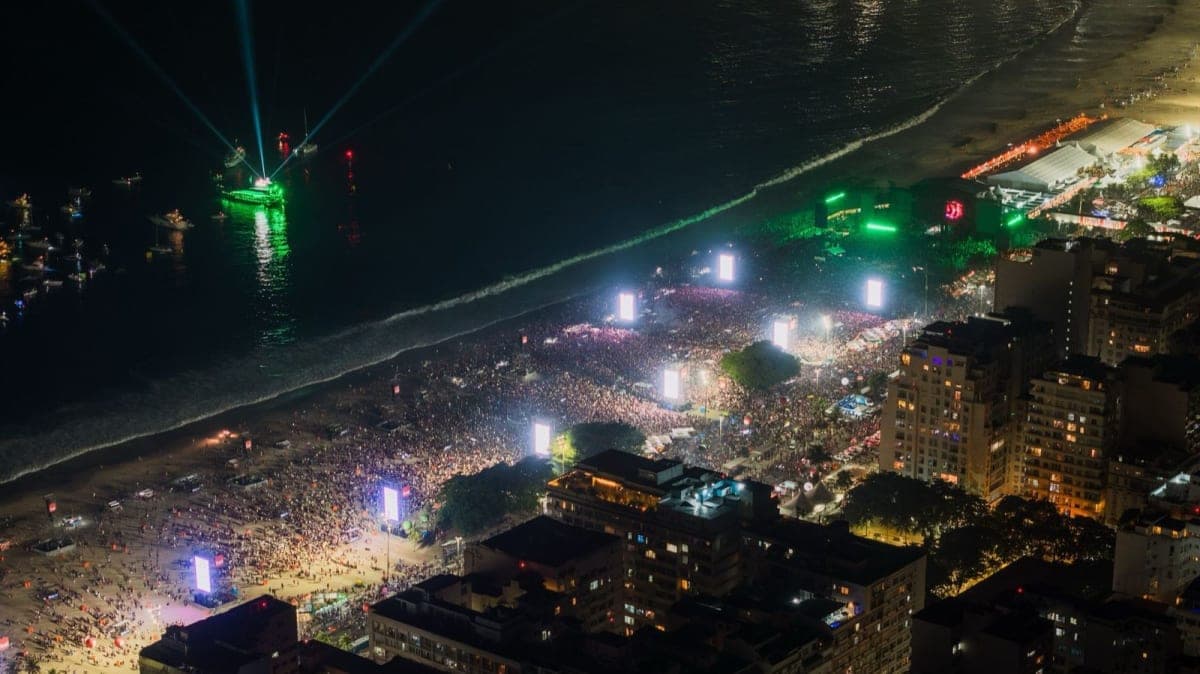 Multitudes llenando la playa de Copacabana de noche alrededor de los escenarios de los conciertos, con barcos iluminados mar adentro, Rio de Janeiro